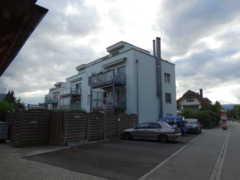 Modern multi-story building with balconies, surrounded by a wooden fence and parked cars.