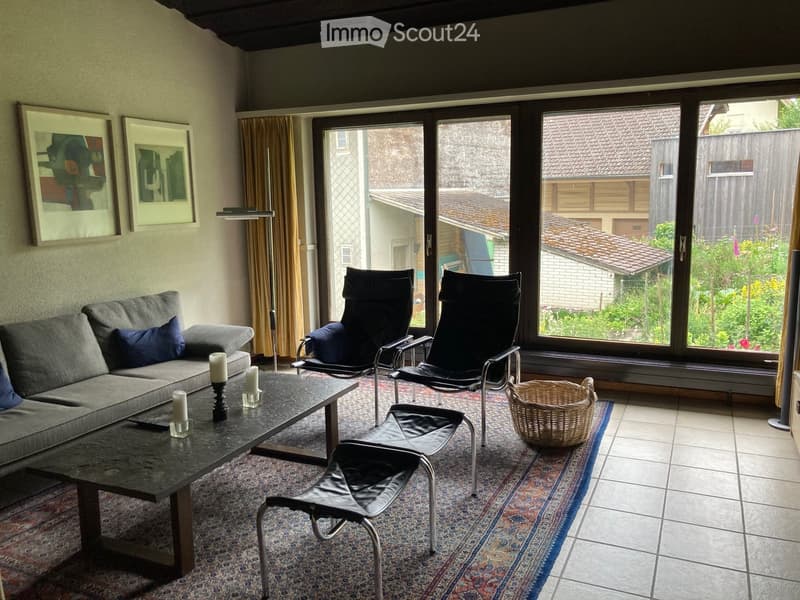 Living room with a grey sofa, coffee table, chairs, a wicker basket, and candles. Large windows offer a view of the garden.