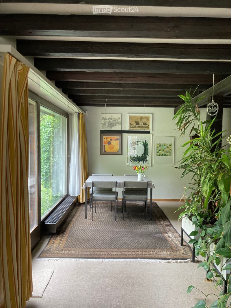 Bright dining area with a wooden ceiling, large windows, and modern artwork on the walls. Includes a dining table with chairs, a patterned rug, and a potted plant.