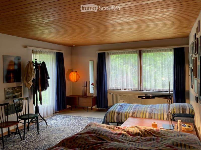 Cozy bedroom with wooden ceiling, large windows with curtains, bed with colorful bedding, wooden chair, small table, lamp, mirror, and coat rack.