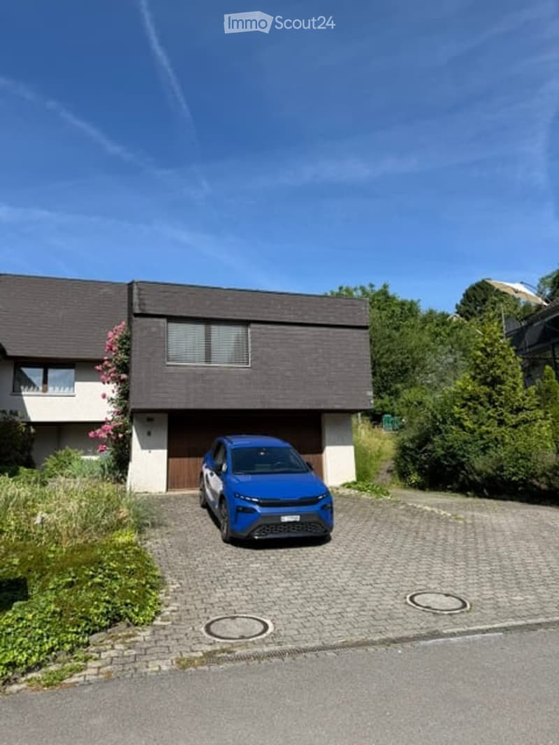 Modern two-story house with a dark roof and white walls, featuring a garage and a parking area with a blue car.