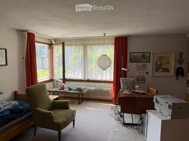 A furnished room with a bed, a green armchair, a desk with a computer, a printer, and a small table with books and a lamp. The room has large windows with red curtains and several picture frames on the wall.