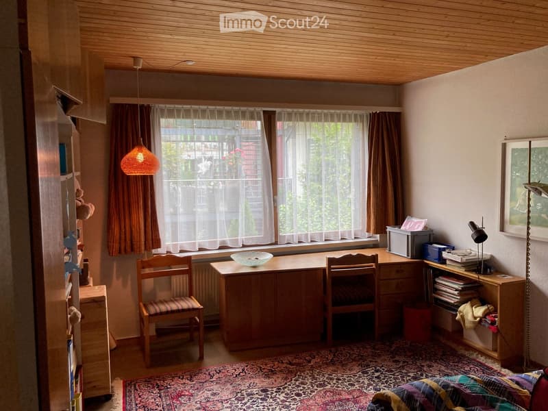 Cozy room with wooden furniture, a bed, a desk, chairs, a lamp, and a bookshelf. Large windows with curtains let in natural light.