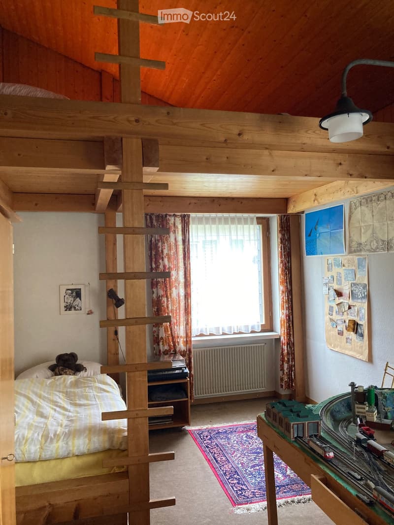 Cozy attic bedroom with wooden beams, a loft bed, a toy train set on a table, and a colorful rug on the floor.