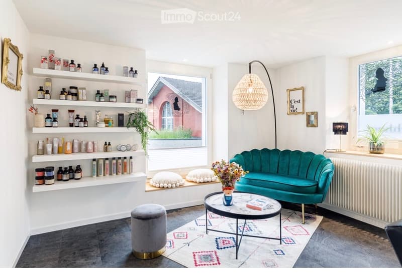 A stylish, well-lit room with a teal sofa, a round coffee table, a grey stool, and shelves filled with various bottles and containers. The room features a large window with a view, a woven pendant light, and decorative elements like a framed picture and a