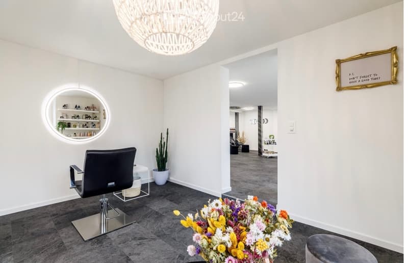 Modern, well-lit room with a black salon chair, a small round table, a flower arrangement, a shelf with various items, and a decorative framed sign on the wall.