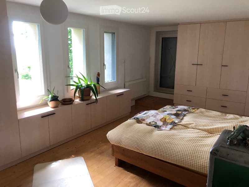 Bright bedroom with wooden flooring, large windows, a double bed with a checkered duvet, built-in wardrobe, and a small table with potted plants.