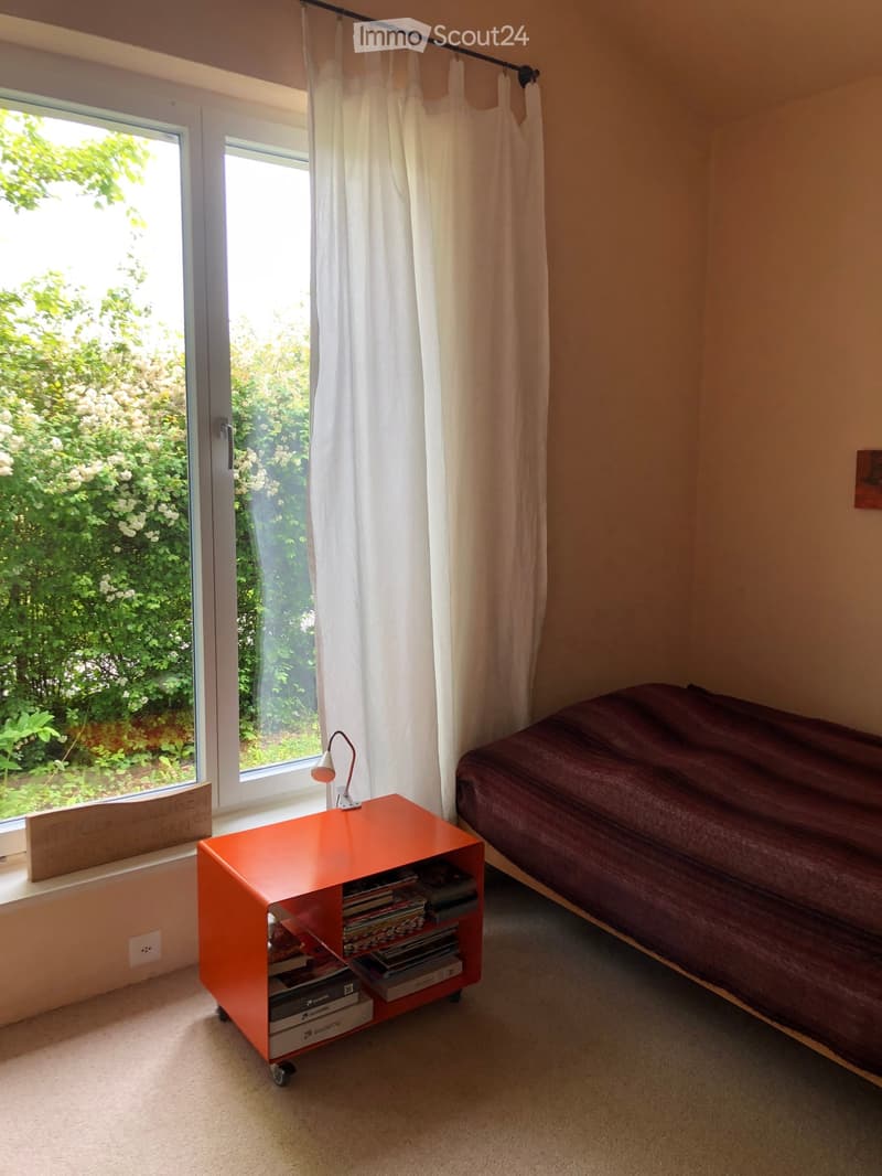 Bright bedroom with a bed, nightstand, lamp, and books. Large window with white curtains offers a view of greenery outside.