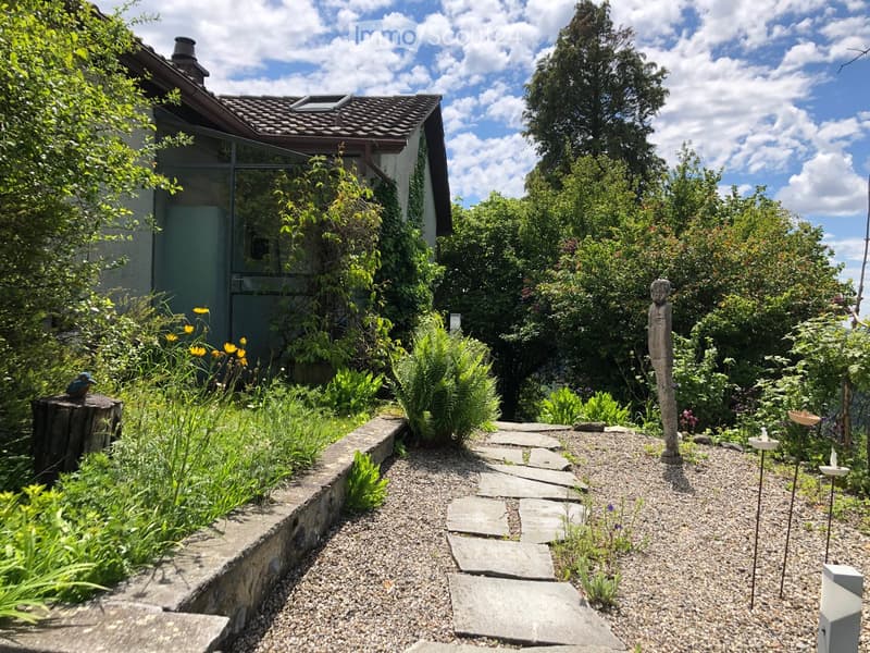Garden with a stone pathway, lush greenery, and a small birdbath. A wooden bird perch and a bird statue are visible.
