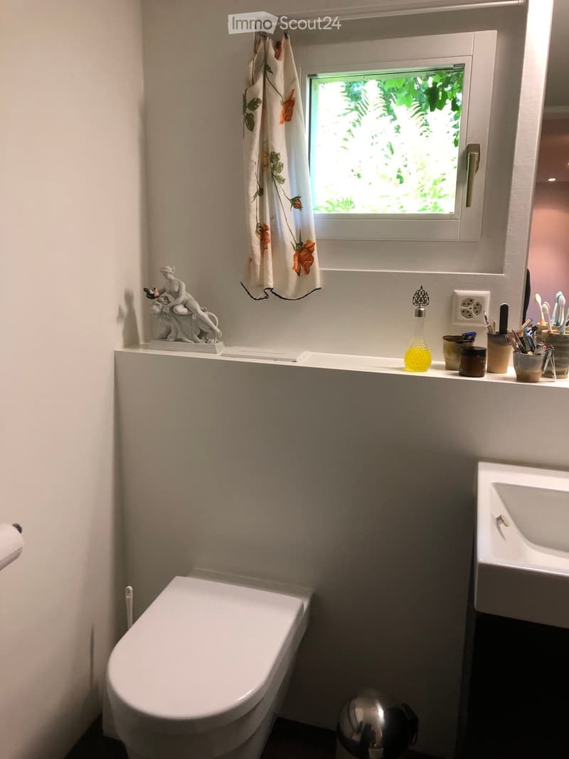 A modern bathroom with a toilet, sink, bathtub, and various toiletries on the counter. A decorative statue and a floral curtain add a touch of elegance.