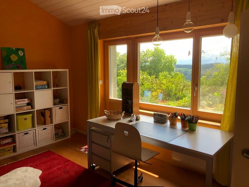 Bright room with large windows offering a scenic view, furnished with a desk, chair, shelves, and decorative items.