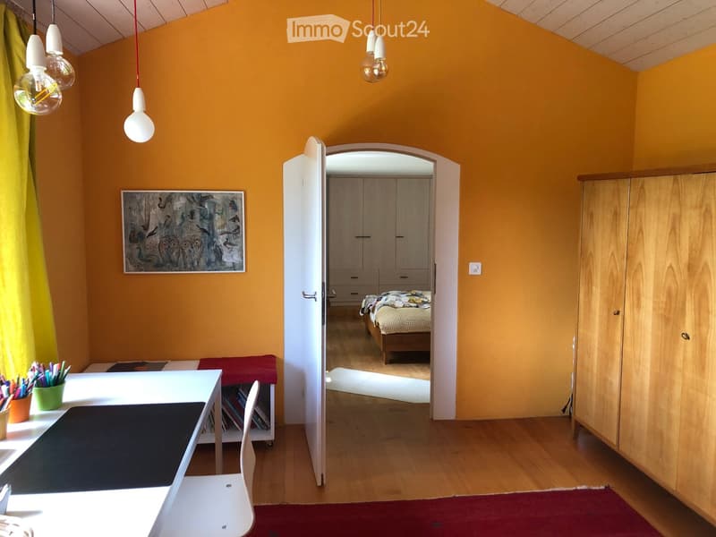 Bright room with wooden flooring, a white table with a chair, a red rug, a large cabinet, and a doorway leading to a bedroom with a bed.
