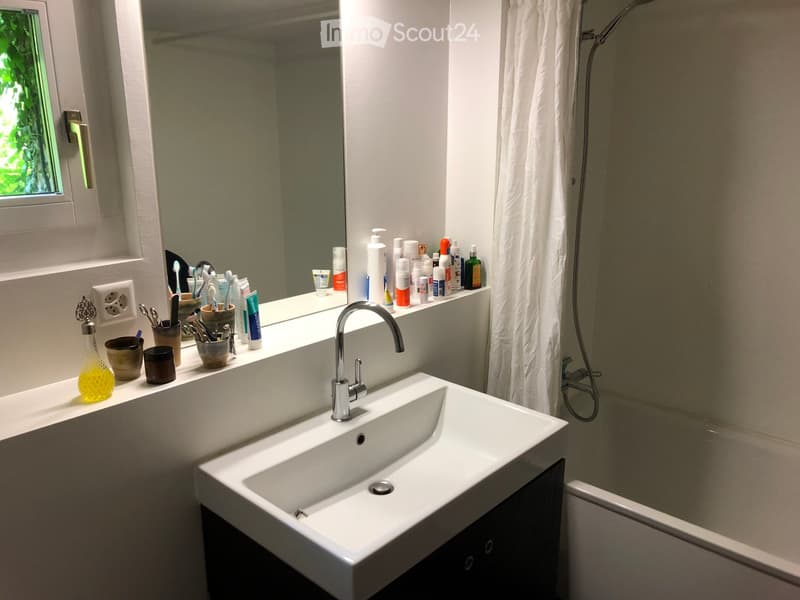 Modern bathroom with a white sink, chrome faucet, large mirror, bathtub, and shower curtain. The countertop holds various toiletries, including toothbrushes, toothpaste, and cosmetics.