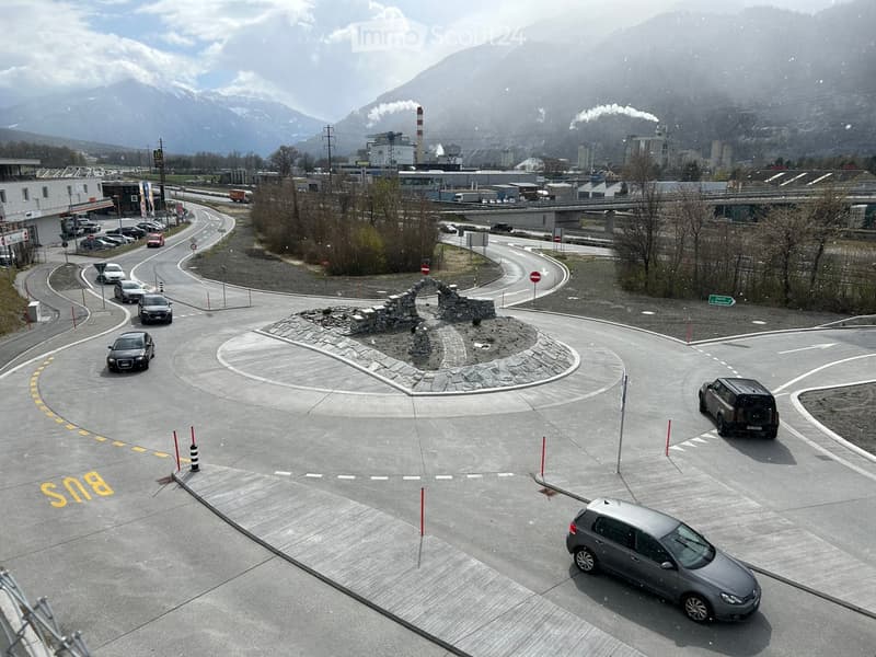 A roundabout with several cars navigating it, surrounded by industrial buildings and a mountainous backdrop.