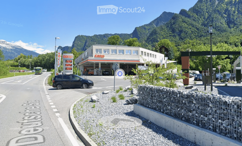 A modern gas station building with a sign displaying fuel prices, located on a road with cars passing by, surrounded by greenery and mountains.