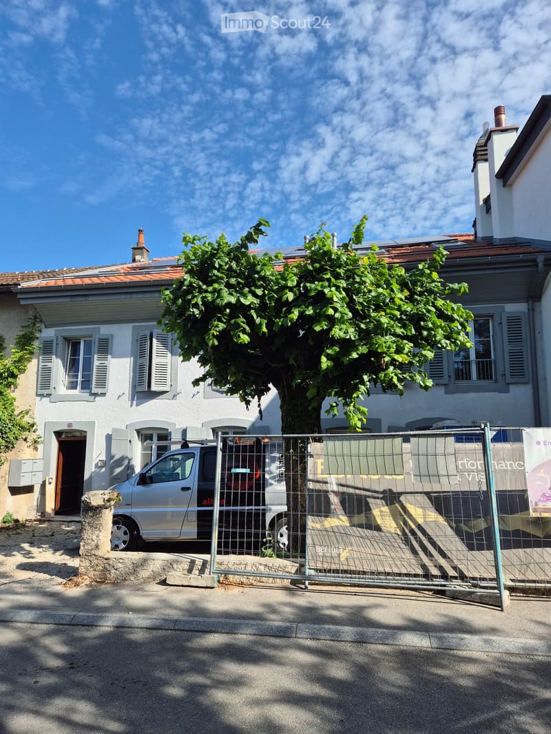 Two-story house with white walls, grey shutters, and a red-tiled roof. A large tree shades the front yard, and a silver van is parked behind a temporary fence.
