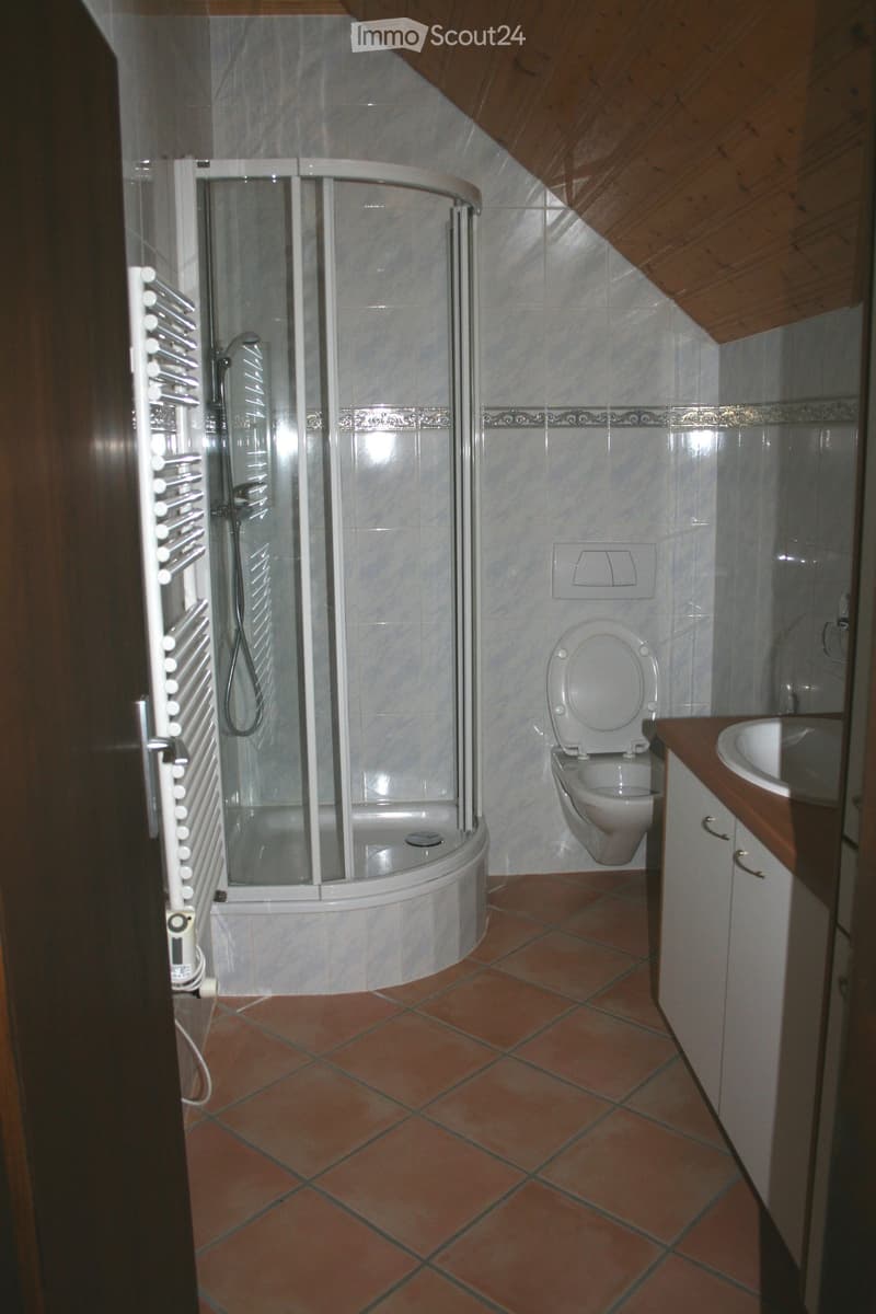 Bathroom with a corner shower, toilet, sink, and radiator. The walls are tiled in a light marble pattern, and the floor is covered with terracotta tiles.