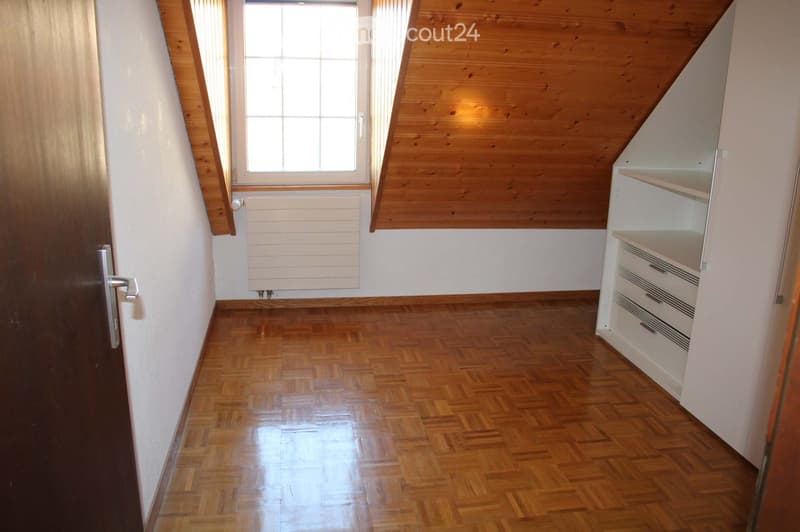 Empty attic room with wooden slanted ceiling, wooden floor, white walls, window, radiator, and built-in white shelving unit with drawers.