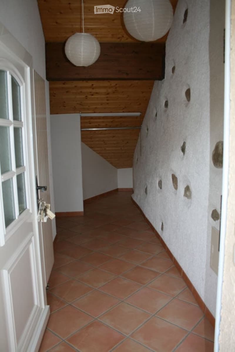 A narrow hallway with exposed wooden beams on the ceiling, terracotta tiled floor, and white walls with decorative stone accents. Two hanging spherical lamps provide lighting.