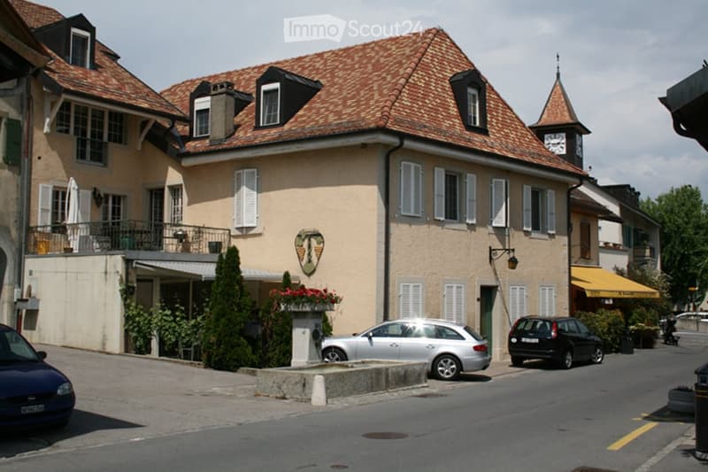 A multi-story building with a tiled roof, white shutters, and a clock tower. The building has a beige facade and is surrounded by parked cars and greenery.