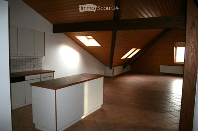 An attic space with exposed wooden beams, skylights, and a small kitchen area. The room is empty, with no furniture or personal items.
