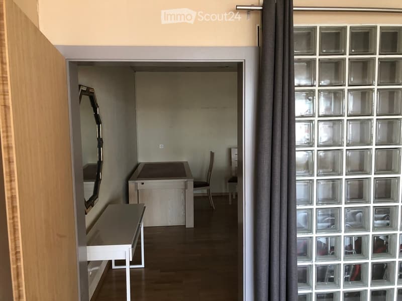 A small, furnished room with a wooden floor, a mirror on the wall, a small table, and a chair. The room is connected to a hallway with a glass-block wall and a curtain.