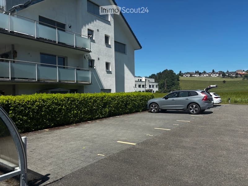 Modern multi-story apartment building with balconies, parked cars in the foreground, and a well-maintained hedge lining the driveway.