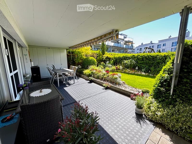Balcony with outdoor furniture, including a wicker chair, a small table, and a dining set, overlooking a well-maintained garden with green lawns, shrubs, and colorful flowers.