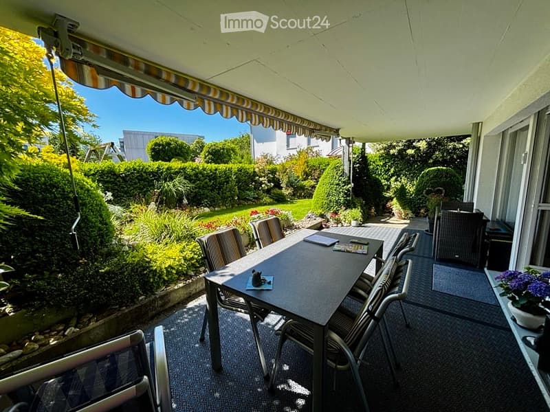 A covered balcony with a dining table and chairs, overlooking a well-maintained garden with various plants and trees.