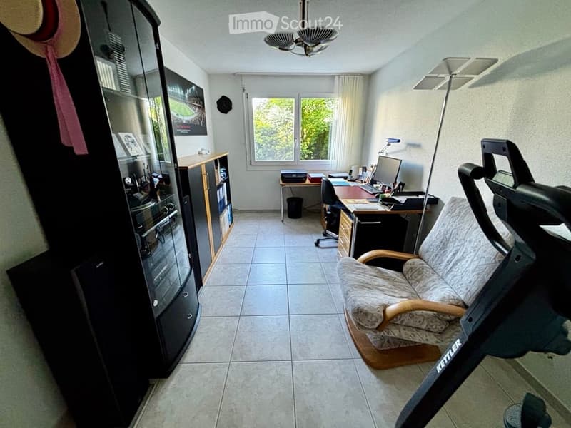 A well-lit room with a sofa, desk, chair, cabinet, mirror, and a lamp. The room features a tiled floor and large windows with curtains.
