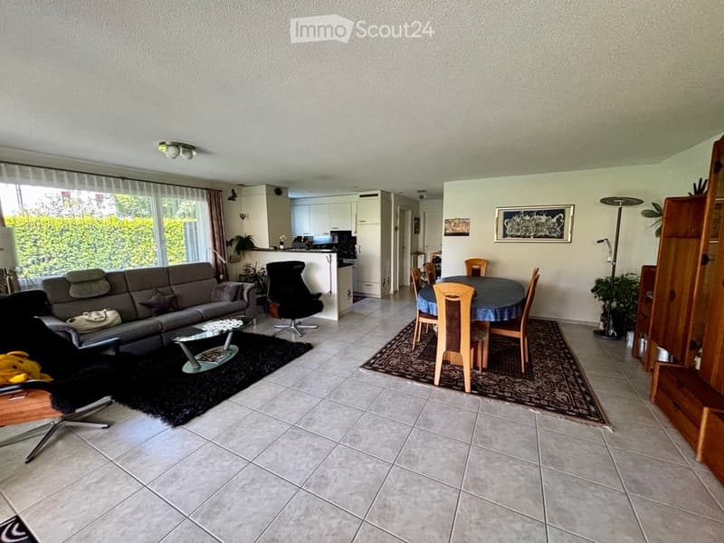 Open-plan living and dining area with a grey sofa, coffee table, dining table with chairs, and a modern kitchen area in the background.