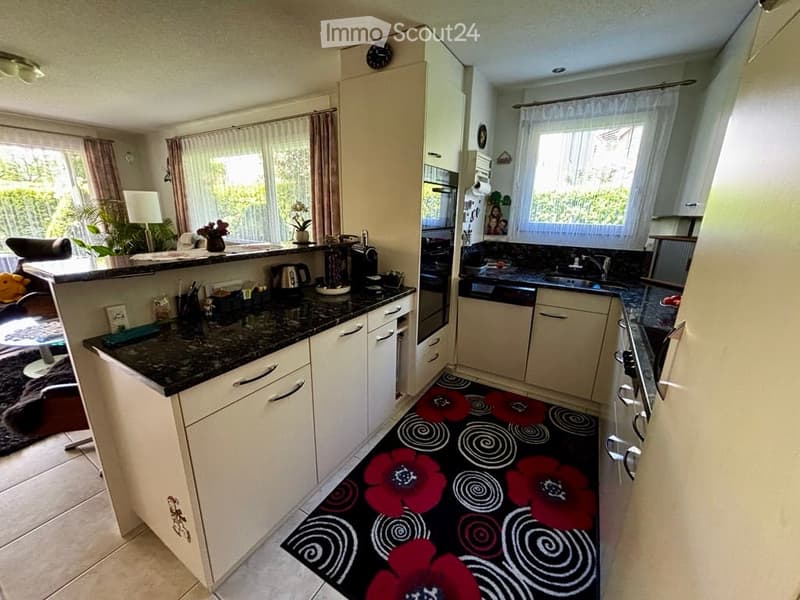 Bright kitchen with white cabinets, granite countertops, and a black and red floral rug. Features include a sink, oven, fridge, coffee maker, kettle, and a small seating area with a chair and table.