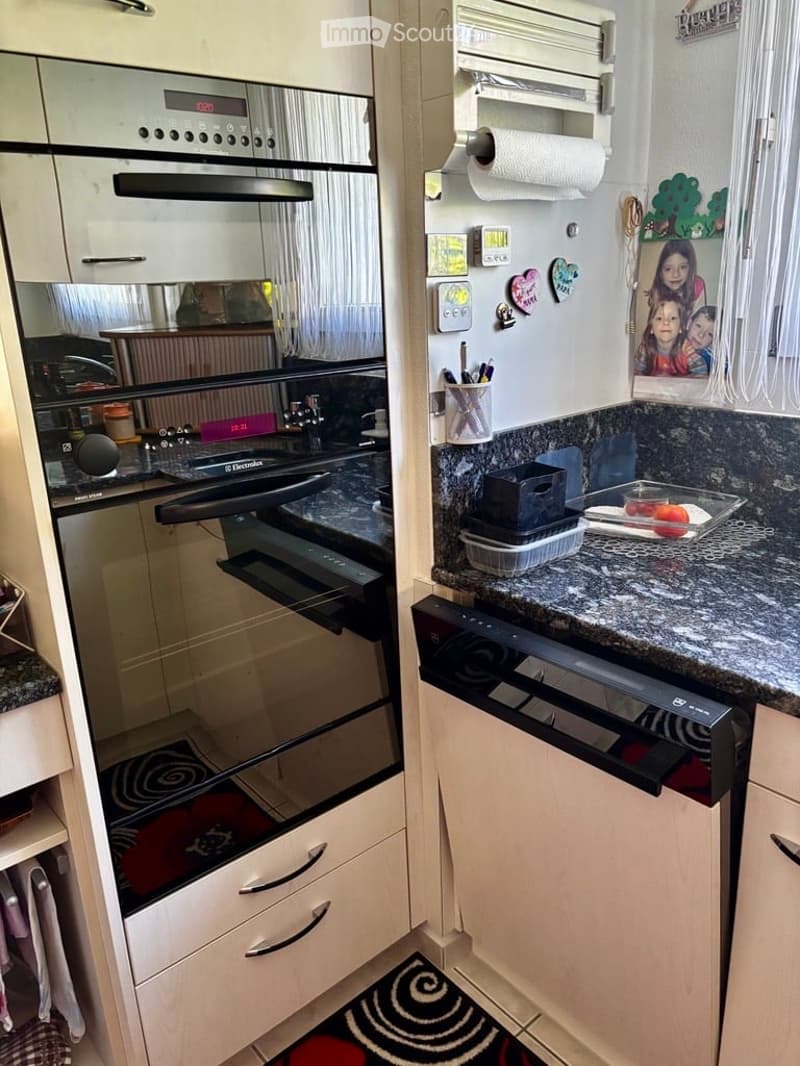 Modern kitchen with white cabinets, granite countertops, built-in oven, refrigerator, and dishwasher. Features include a paper towel holder, small decorative magnets, and a picture frame on the fridge.
