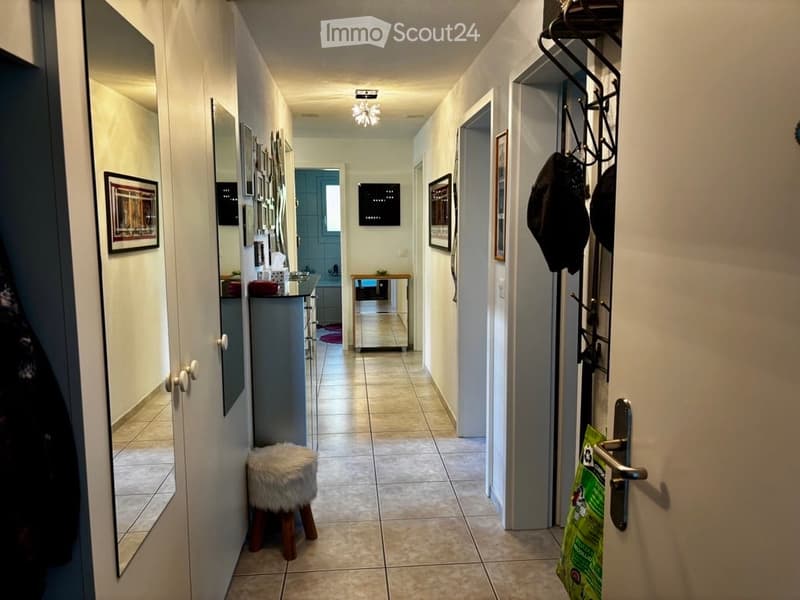 A well-lit hallway with tiled flooring, white walls, and several doors leading to different rooms. There are framed pictures on the walls, a small wooden stool with a fur cushion, and a coat rack with jackets hanging on it.