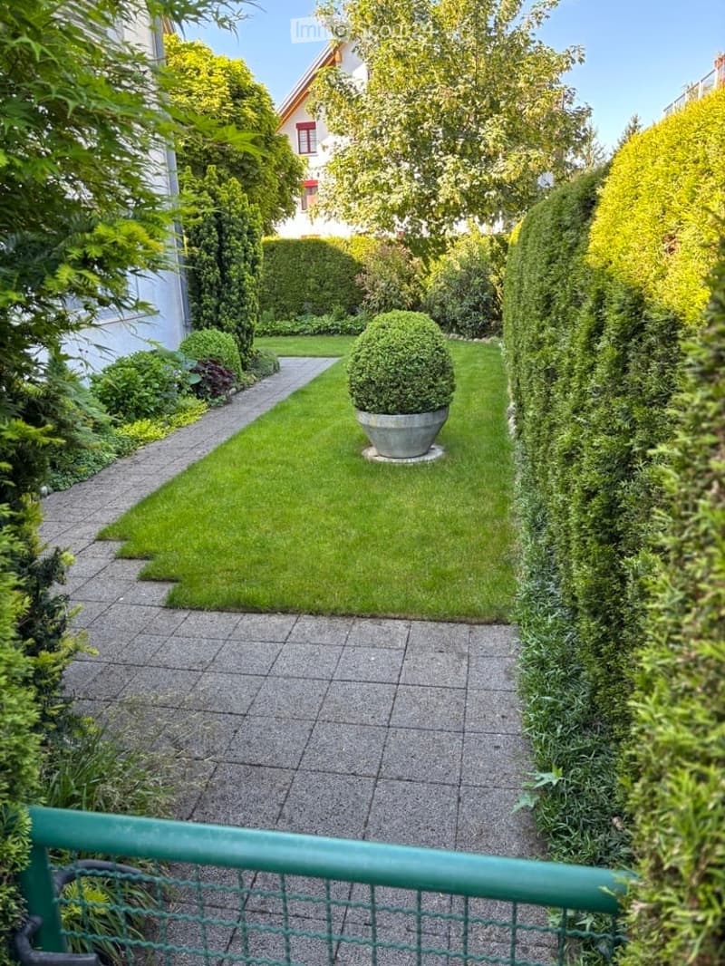 Well-maintained garden with a central lawn, trimmed hedges, and a large potted plant. The garden features a paved pathway leading to the house.