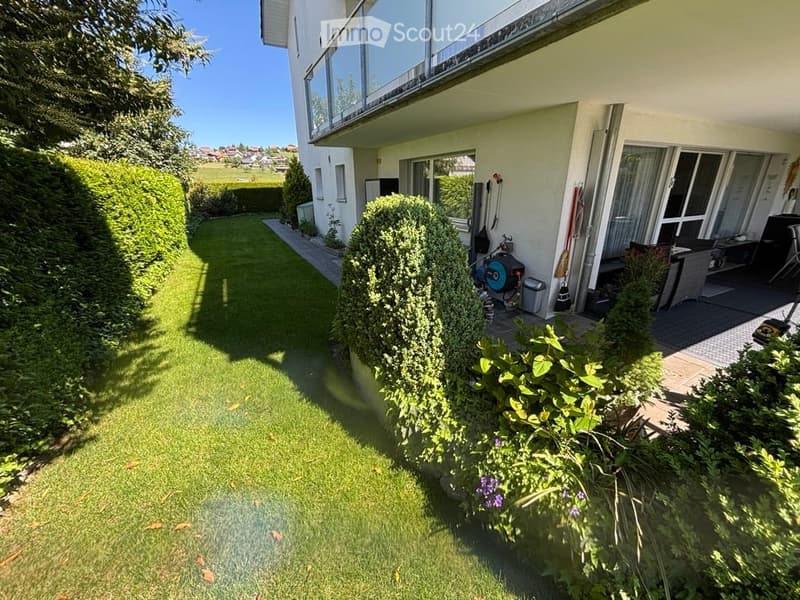 Well-maintained garden with neatly trimmed hedges, a small patio area with outdoor furniture, and a modern house with large windows and a balcony.