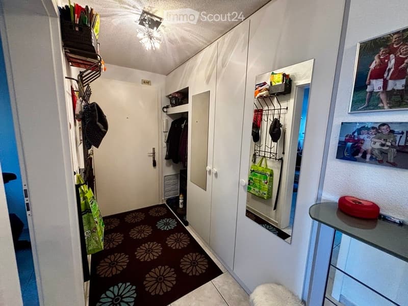 A narrow hallway with white walls, a patterned carpet, built-in wardrobes, and a small table with a red object on it. The hallway features a closed door and a coat rack with bags and jackets.