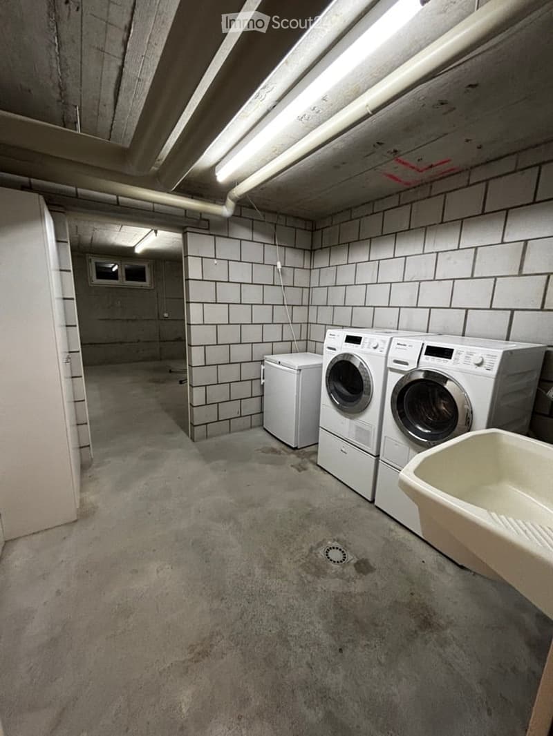 Underground laundry room with concrete floor, tiled walls, fluorescent lighting, and a small window. Contains a washing machine, dryer, and a sink.