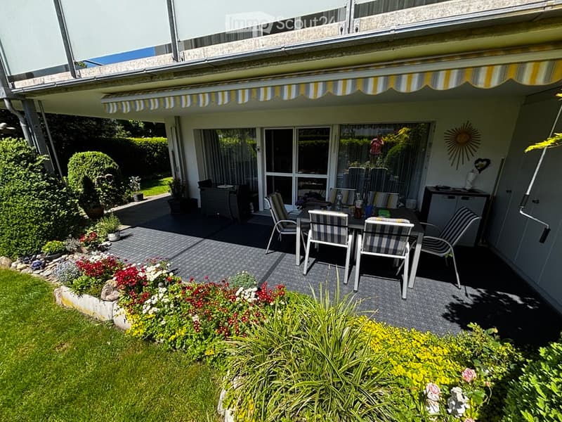 Outdoor patio area with a dining set, striped chairs, and a table, surrounded by a well-maintained garden with colorful flowers and shrubs.