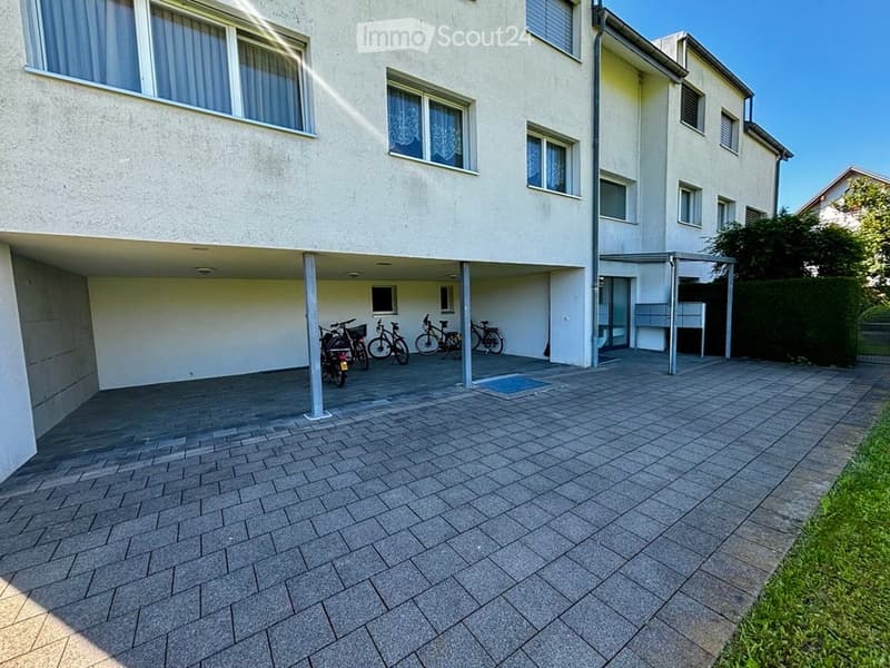 Multi-story building with a covered parking area, several bicycles, and a paved courtyard.