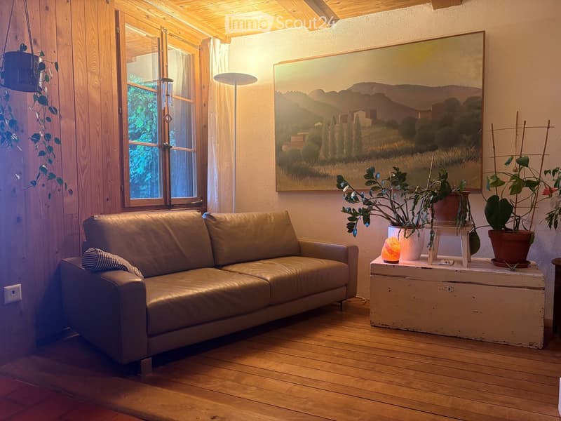 Cozy living room with wooden floors, a large window, a beige leather sofa, a small table with potted plants, and a scenic landscape painting on the wall.