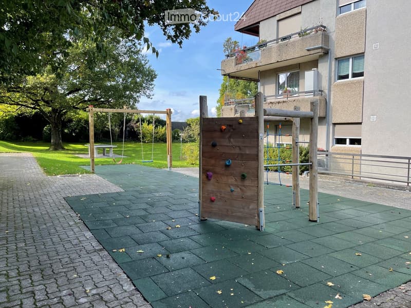 Outdoor playground area with climbing wall, swings, and a small table, adjacent to a modern apartment building with a balcony.
