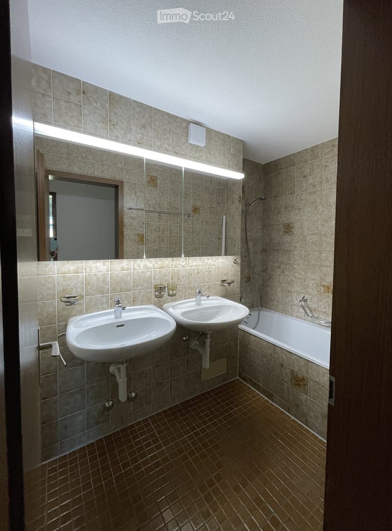 Bathroom with two sinks, a bathtub, a large mirror with built-in lighting, and tiled walls and floor.