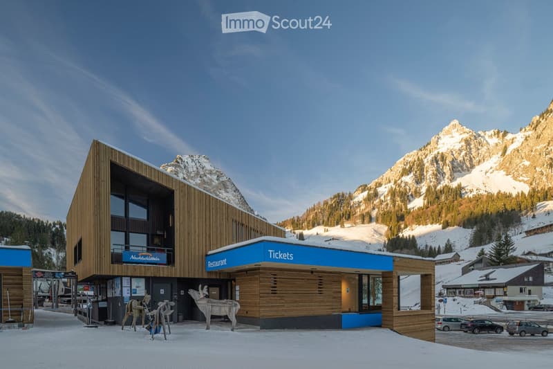 Modern wooden building with a restaurant and ticket office, surrounded by snow-covered mountains and parked cars.