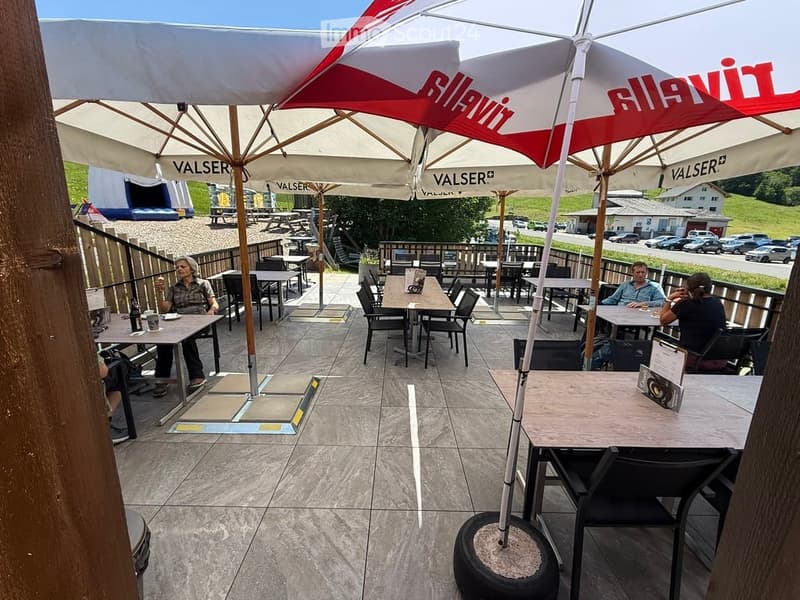An outdoor seating area with tables and chairs under large umbrellas, surrounded by a wooden fence and a view of a parking lot and greenery.