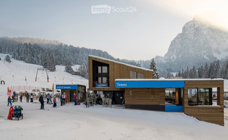 A modern wooden building with a blue ticket booth and a grill & snacks area, surrounded by snowy mountains and a ski slope with people and equipment.