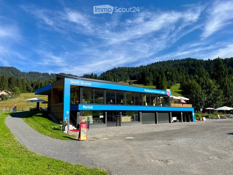 Modern building with a blue facade labeled "Skilift Bruni" and "Restaurant Bruni-Lodge." Features large windows, a covered outdoor seating area with umbrellas, and a gravel driveway.