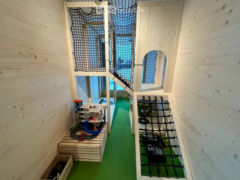 A small indoor play area with wooden walls, green flooring, and a toy car track setup. The space includes a netted climbing area and a small toy house.