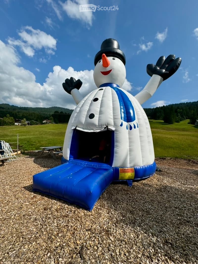 Inflatable snowman bouncy house with slide, placed on a gravel surface in a grassy field with picnic tables and playground equipment nearby.