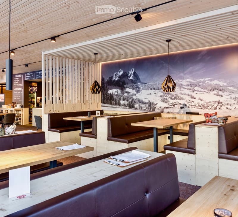 Modern restaurant interior with wooden tables, leather sofas, pendant lights, and a large mural of a snowy mountain landscape.